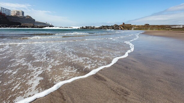 Une plage avec des vagues, une côte rocheuse et des bâtiments en arrière-plan.
