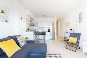 A modern living room with a sofa, a dining area with a table and chairs, a kitchen with white cabinets and appliances, and a small office space.