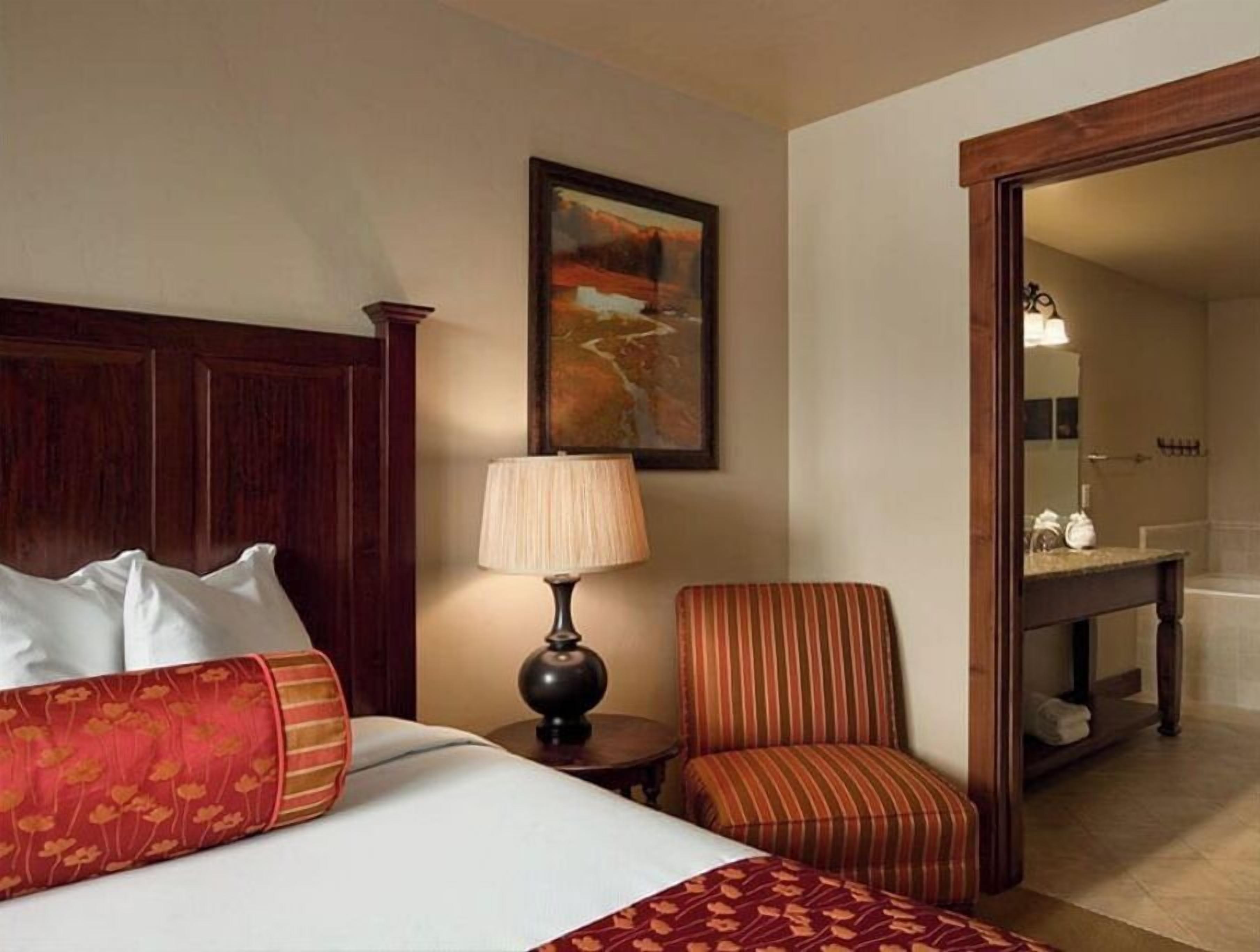 Habitación de hotel con una cama grande, una mesita de noche con lámpara, un sillón a rayas, un cuadro en la pared y un espejo que refleja un baño.