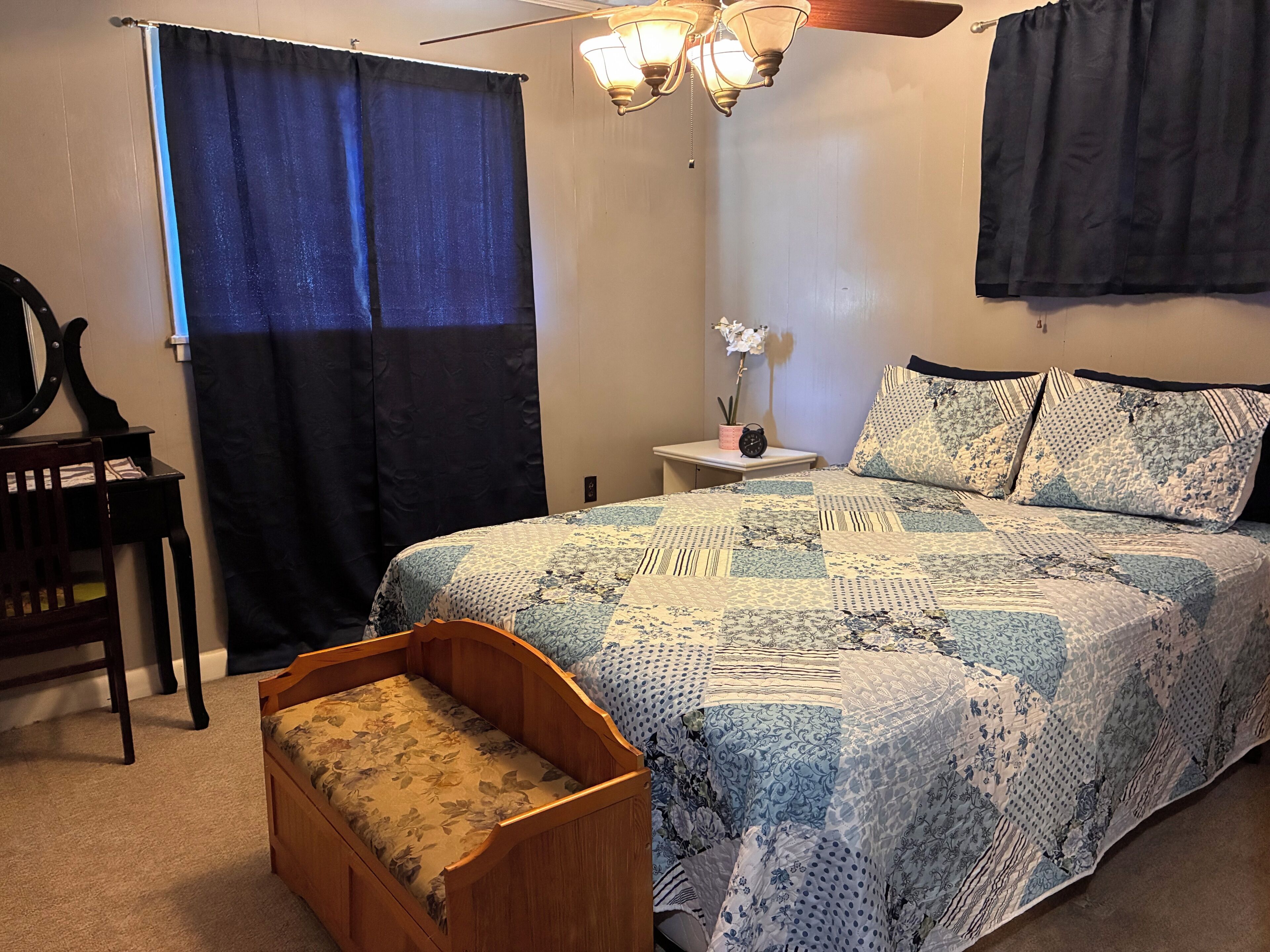 Une chambre à coucher comprenant un lit, une table de chevet, une chaise, une fenêtre avec des rideaux et un ventilateur de plafond.