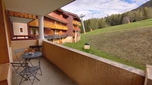 A balcony with a table and chairs, overlooking a grassy hillside and a building with balconies. - Apartment at the foot of the slopes in a typical village of our mountains (Les Coches, La Plagne-Tarentaise, France)