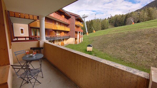 A balcony with a table and chairs, overlooking a grassy hillside and a building with balconies.