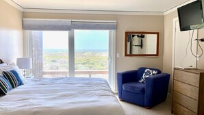 A hotel room with a large bed, a blue armchair, a wooden dresser, and a television. - Serene beachfront getaway, breathtaking ocean views, steps from the beach! (Moss Landing)