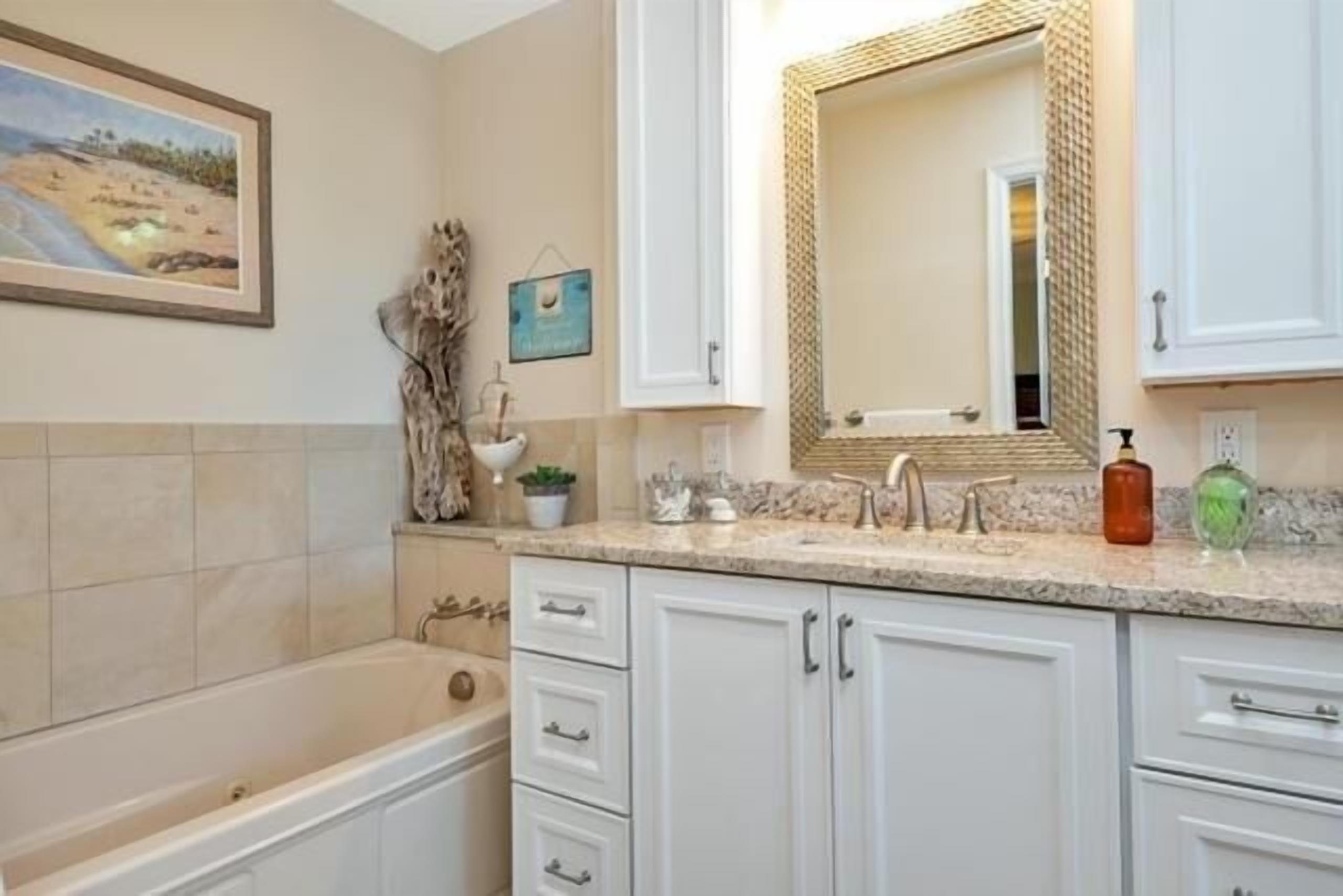A bathroom with a bathtub, white cabinets, a sink with a mirror, and a framed picture on the wall.