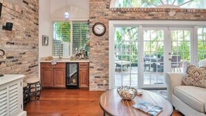 Amplia sala de estar con cocina, armarios de madera, una mesa de centro redonda y un gran ventanal con vista a muebles de exterior.