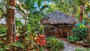 Una cabaña con techo de paja y un sendero de piedra que la conduce, rodeada de exuberante follaje tropical y una valla de madera.