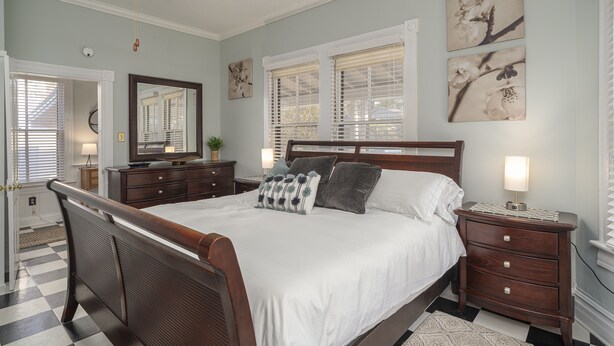 A bedroom with a large bed, a wooden headboard, a nightstand with a lamp, and a dresser. There are two windows with blinds, a ceiling fan, and artwork on the walls.