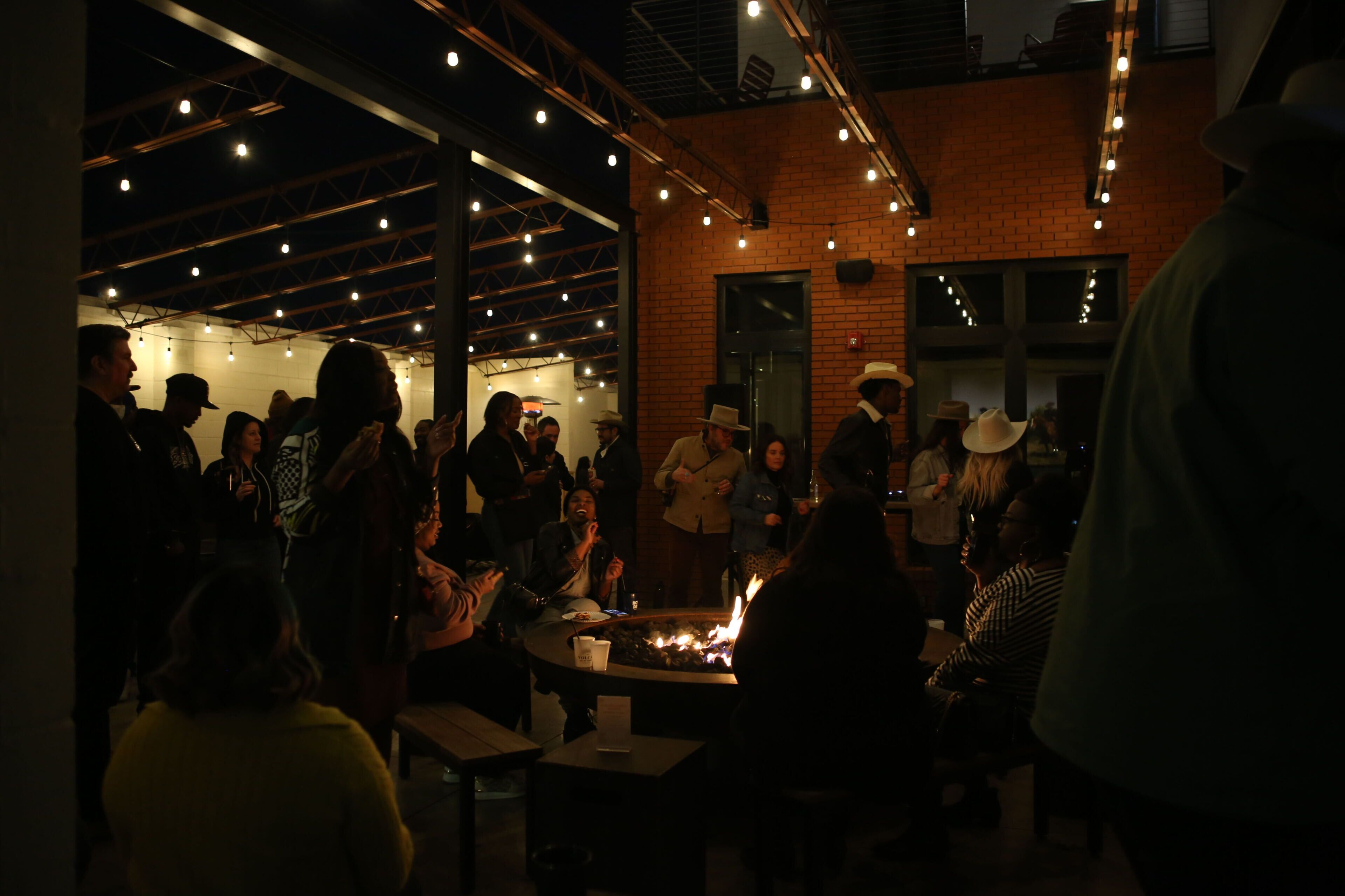 A group of people gathered around a fire pit in a dimly lit indoor setting with brick walls and exposed beams.
