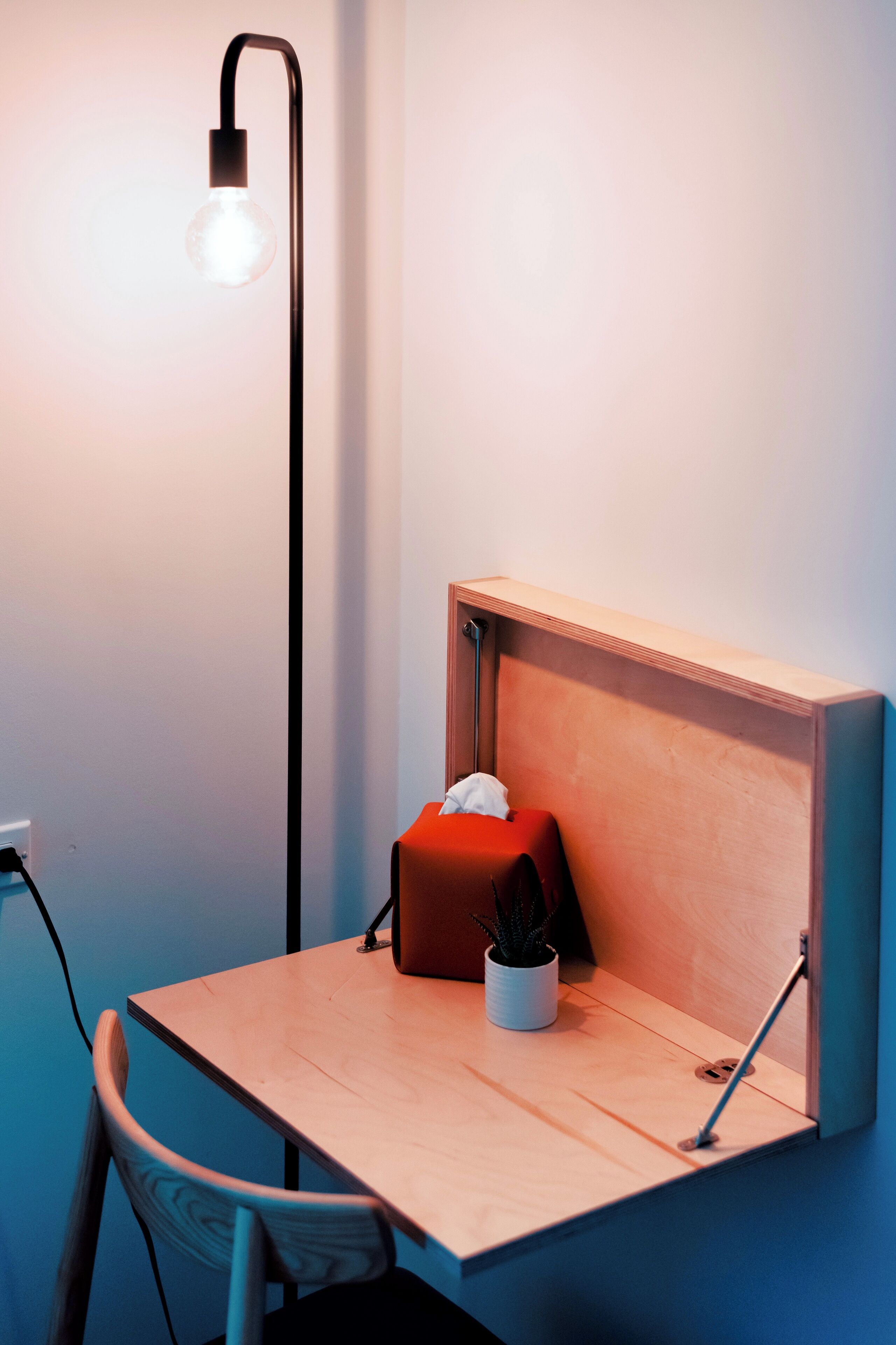 A small wooden desk with a red box and a white pot, a chair, and a tall floor lamp.