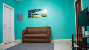 A room with teal walls, a brown sofa, a grey rug, a white door, and a TV on a wooden stand. - Near the beach family (West End)