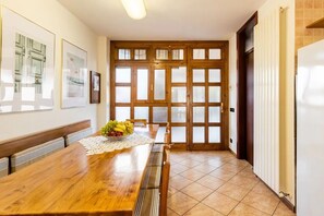 A dining area with a wooden table, chairs, and a fruit bowl. There are framed pictures on the wall, a radiator, and a door with frosted glass panels.