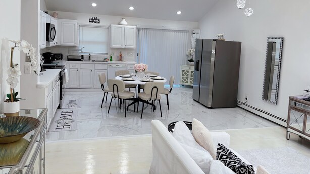 A modern kitchen with white cabinets, a dining area with a round table, and a living area with a sofa and decorative pillows. - Cozy Hamptons Escape 3BR Minutes From Beach! (Hampton Bays)