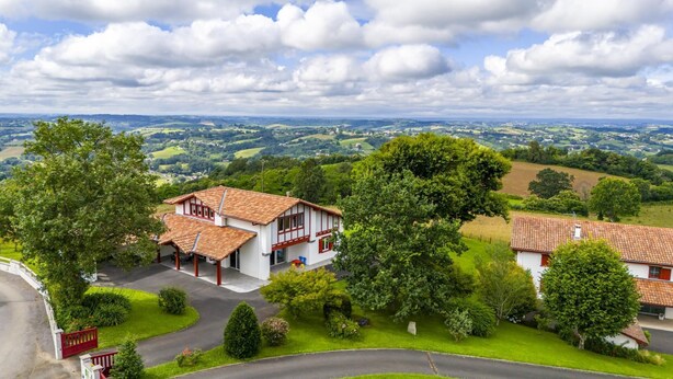 Ein traditionelles Chalet mit geneigtem Dach, umgeben von üppiger Vegetation und einem malerischen Ausblick auf sanfte Hügel.