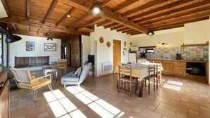 Amplia sala de estar con techo de madera, una mesa de comedor para cuatro personas y una cocina al fondo.