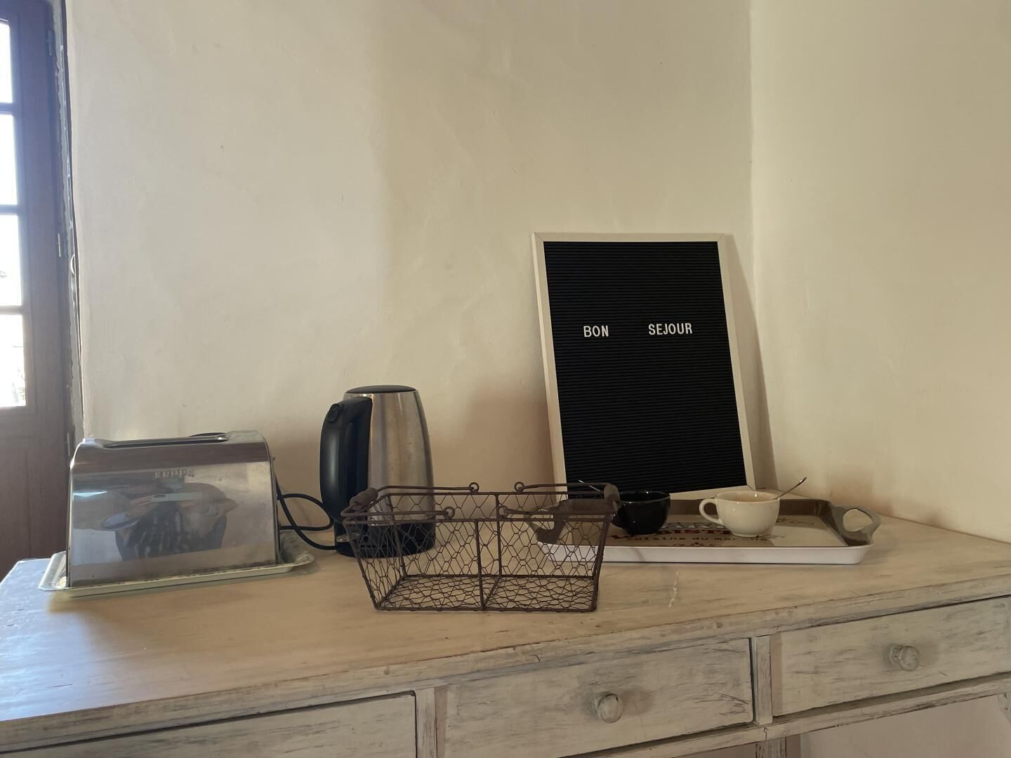 A kitchen counter with a coffee maker, a kettle, a tray, and a sign that reads "BON SEJOUR".