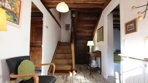 Eine Holztreppe führt zu einer zweiten Etage, ein Sessel mit Kissen, ein Tisch mit Lampe und eine Wanduhr.