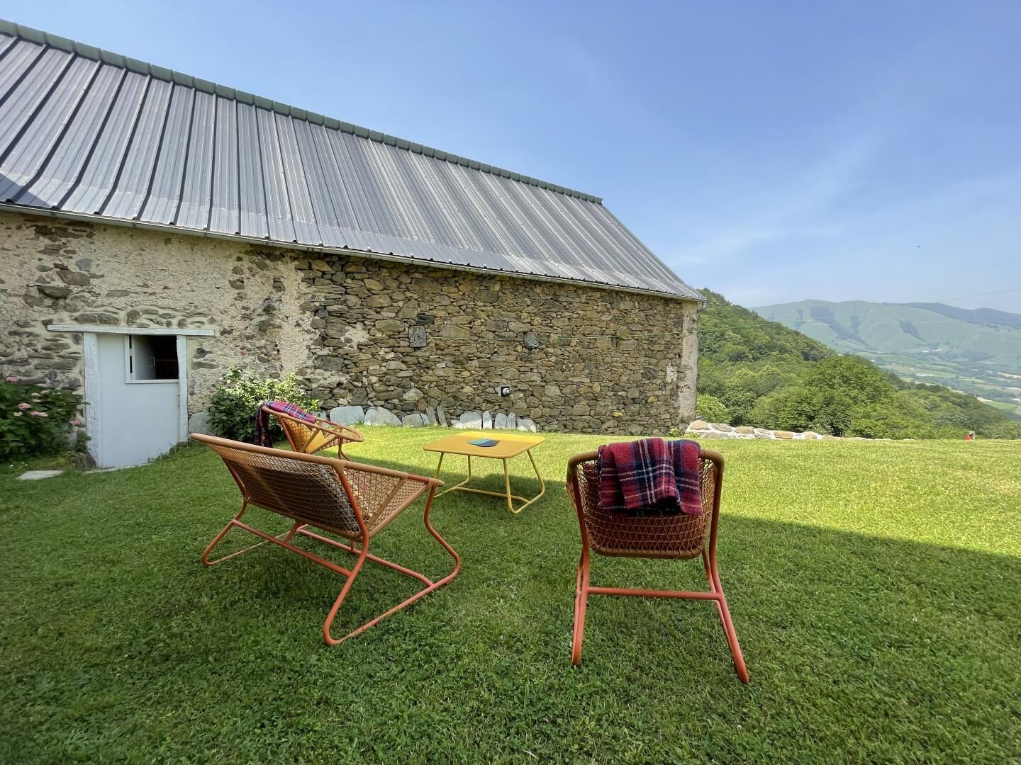 A stone building with a metal roof, a small door, and a grassy lawn with two wicker chairs and a yellow table.