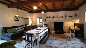 Ein gemütliches Wohnzimmer mit Holzbalkendecke, Kamin, einer Couch, einem Esstisch mit Stühlen und einer Bank.