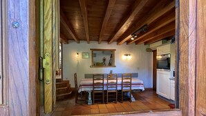 Un coin repas rustique avec une table et des chaises en bois, un sol carrelé et une cuisine visible à travers une porte ouverte.