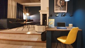 Een moderne woonkamer met een donkerblauwe muur, een gele fauteuil, een blauwe bank, een houten bureau met een laptop en een lijst met een foto aan de muur.