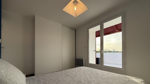 A bedroom with a bed, a wardrobe, a radiator, and a window with a view of a parking area. - Xokobia - Arbonne - Gîte (ARBONNE)