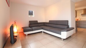 A modern living room with a grey sectional sofa, a flat-screen TV on a wooden stand, and a small window. - Xokobia - Arbonne - Gîte (ARBONNE)