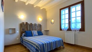 Ein Schlafzimmer mit einem hölzernen Kopfteil, einem Bett mit blauer Bettwäsche, zwei Wandlampen, einem Fenster mit weißen Vorhängen und einem Heizkörper.