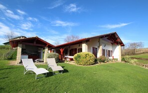 Uma casa de dois andares com telhado vermelho, pilares de pedra e um alpendre com duas poltronas.