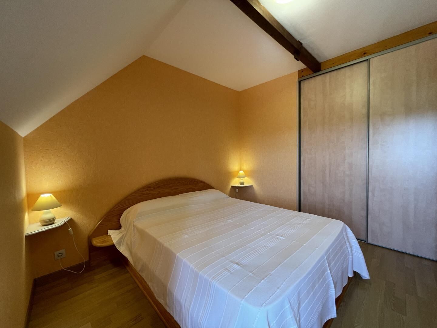 A bedroom with a sloped ceiling, a bed with a white bedspread, two bedside lamps, and a wooden floor.