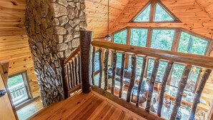 Interior de una cabaña de madera con pared de piedra, techo de madera y un balcón con barandilla.