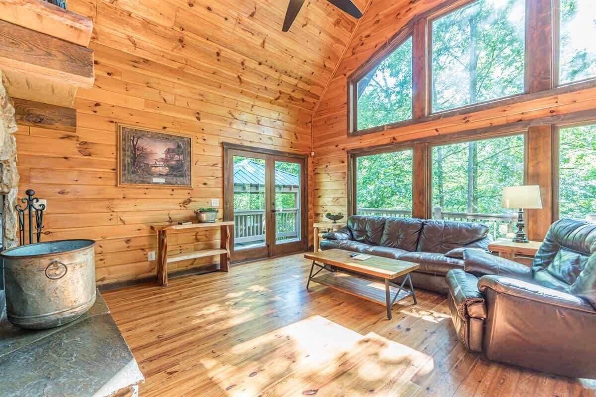 L’intérieur d’un chalet en rondins, avec un plancher en bois, un canapé en cuir, une table basse et de grandes fenêtres donnant sur le paysage extérieur.