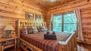 Un dormitorio de cabaña de madera con una cama, almohadas, una mesita de noche, una lámpara y una ventana con cortinas.