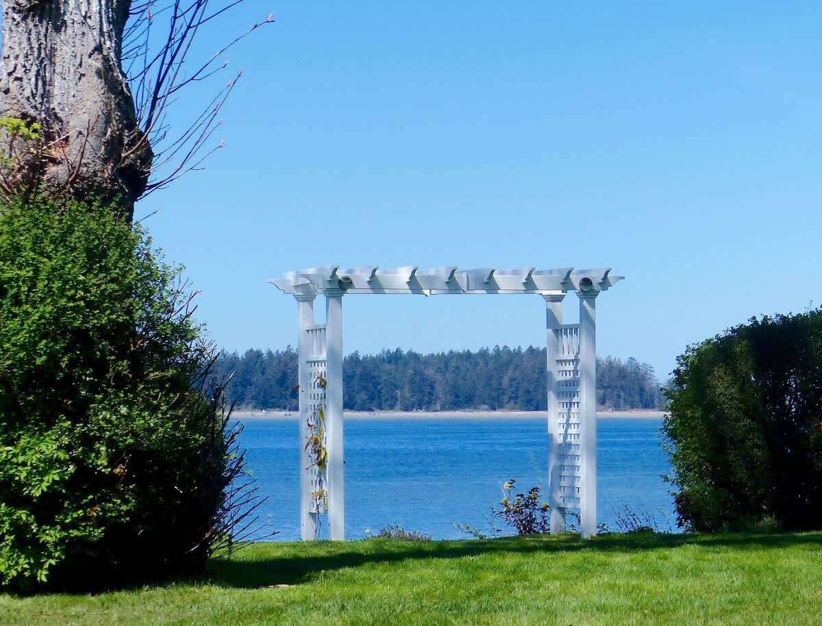 A white pergola with a view of a calm body of water and a forested shoreline.