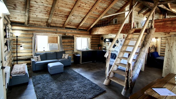 Ein Blockhaus-Innenraum mit Schlafzimmer im Zwischengeschoss, einem Wohnbereich mit Sofa und Hocker, einer Küchenzeile und einer Treppe.