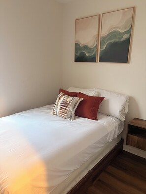 Ein Bett mit weißer Bettwäsche und einem gemusterten Kopfkissen, ein Nachttisch aus Holz sowie zwei abstrakte Gemälde an der Wand.