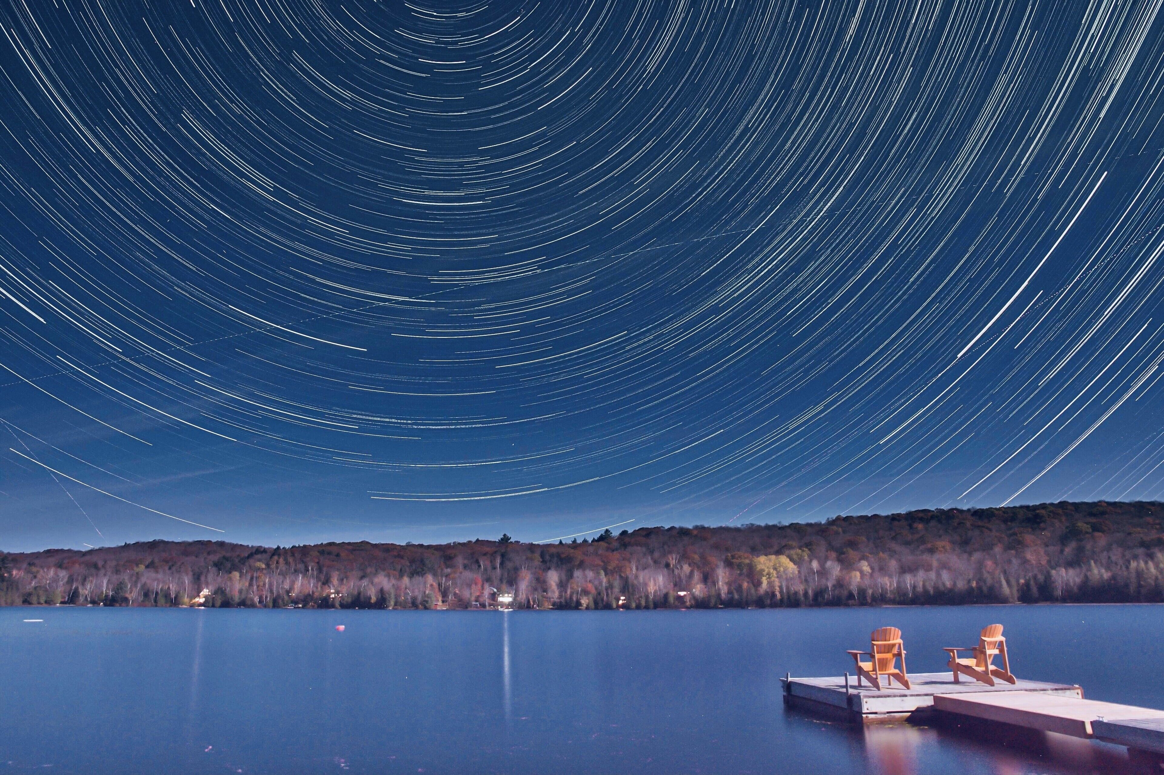 A lake with a star trail effect in the night sky, a dock with two chairs, and a forested shoreline.