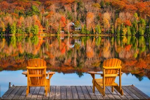 A wooden dock with two Adirondack chairs facing a calm lake, surrounded by autumn-colored trees and a house in the background.