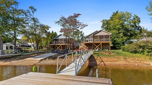 Une jetée en bois menant à une maison dotée d’un balcon, entourée d’arbres et bordée par un plan d’eau.