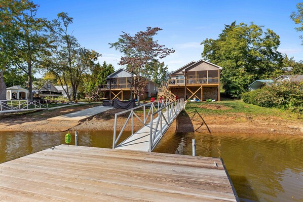 Un muelle de madera que lleva a una casa con balcón, rodeada de árboles y una masa de agua.