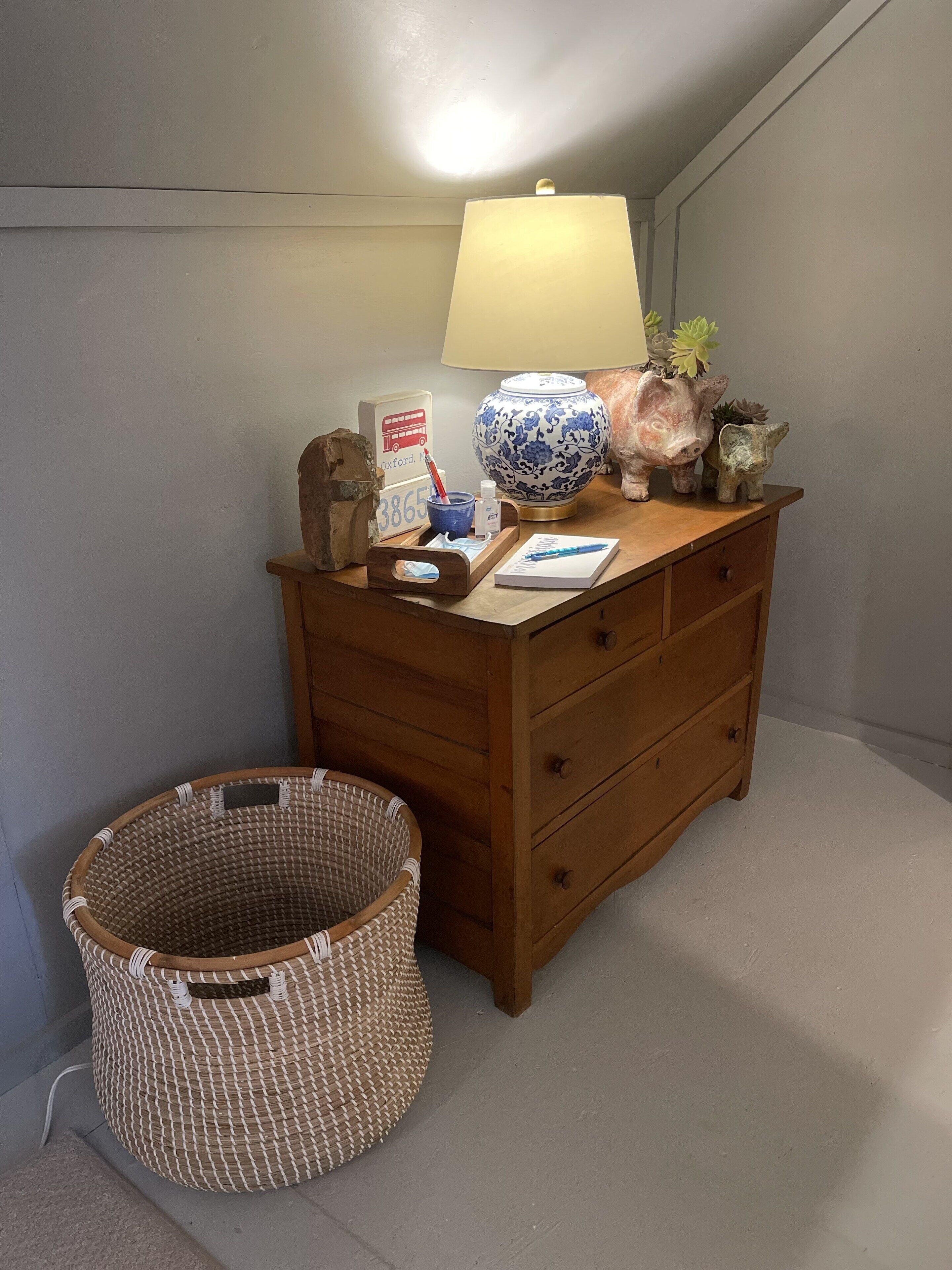 Ein hölzerner Kommode mit einer Lampe, einer Vase und Dekorationselementen. Neben der Kommode steht ein Korbwicker-Korb.