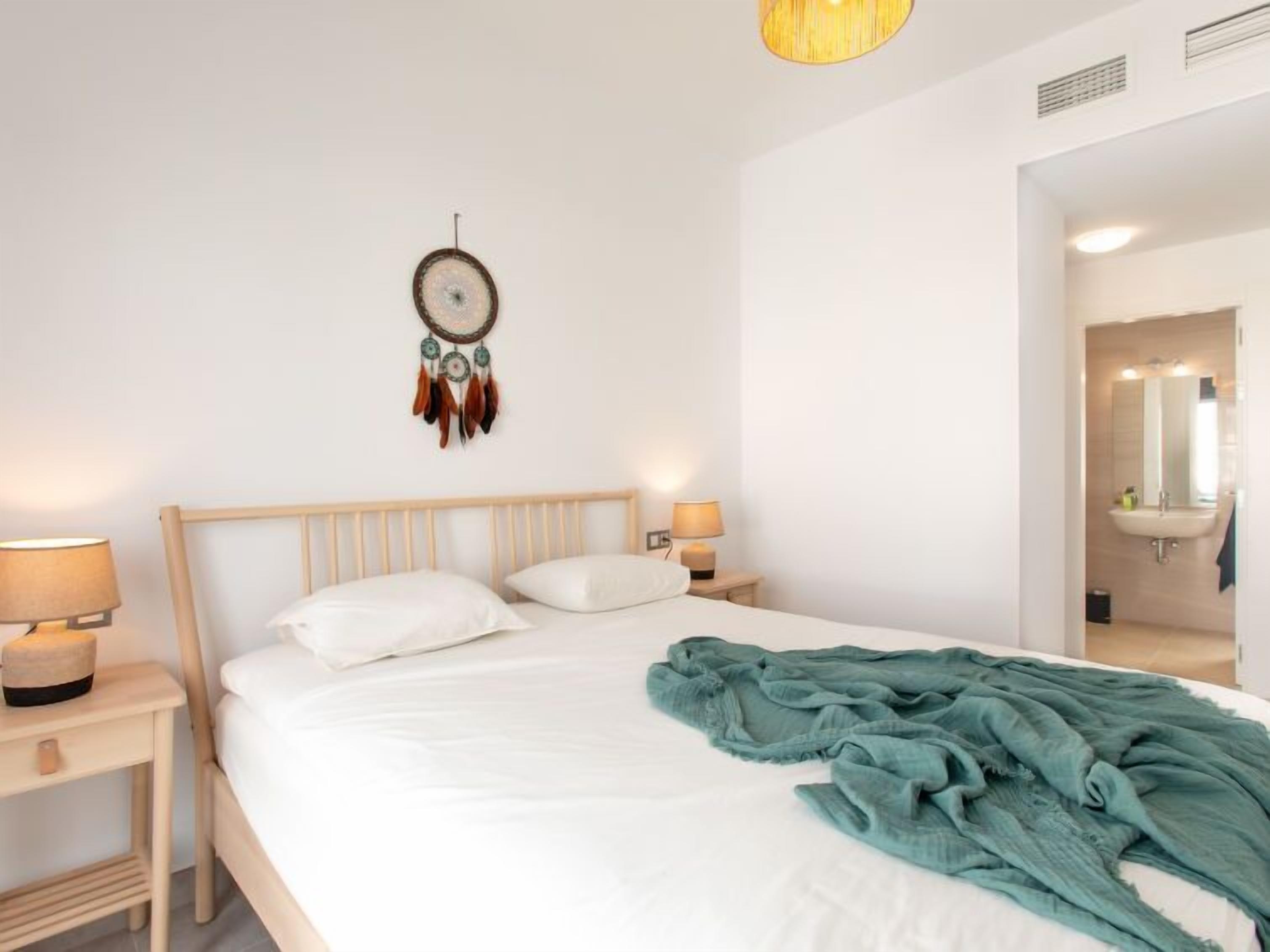 Un lit bien fait, avec des draps blancs, une couverture turquoise et un attrape-rêves accroché au mur.