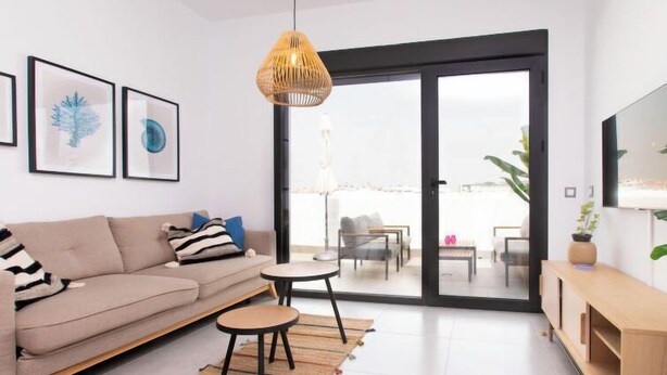 Ein modernes Wohnzimmer mit einer Couch, einem kleinen Tisch und einer Bank. An den Wänden hängen gerahmte Bilder, ein großes Fenster bietet Blick ins Freie.