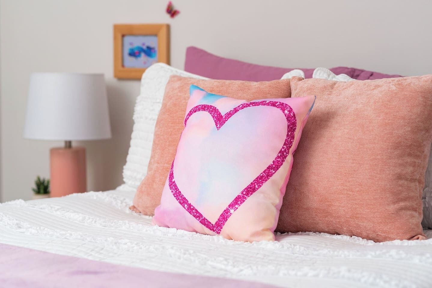 A bed with white bedding, pink and white pillows, a pink lamp, and a framed picture on the wall.