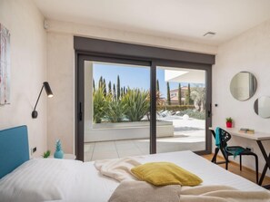 A bedroom with a bed, a desk, and a chair. There is a sliding glass door leading to an outdoor area with plants and a view of houses. - Old Olive I by Interhome (Pula/Vodnjan)