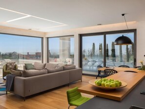 A modern living room with a sofa, a dining table with a fruit bowl, and large windows offering a view of the cityscape. - Old Olive I by Interhome (Pula/Vodnjan)