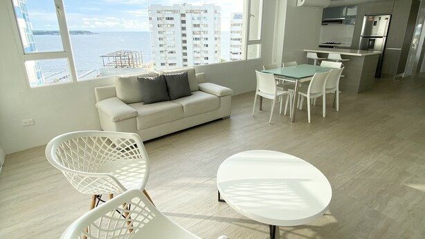 A modern living room with a white sofa, a dining table with chairs, and a view of the sea and buildings. - 3TC10 Apartamento Cartagena frente al mar (Cartagena)