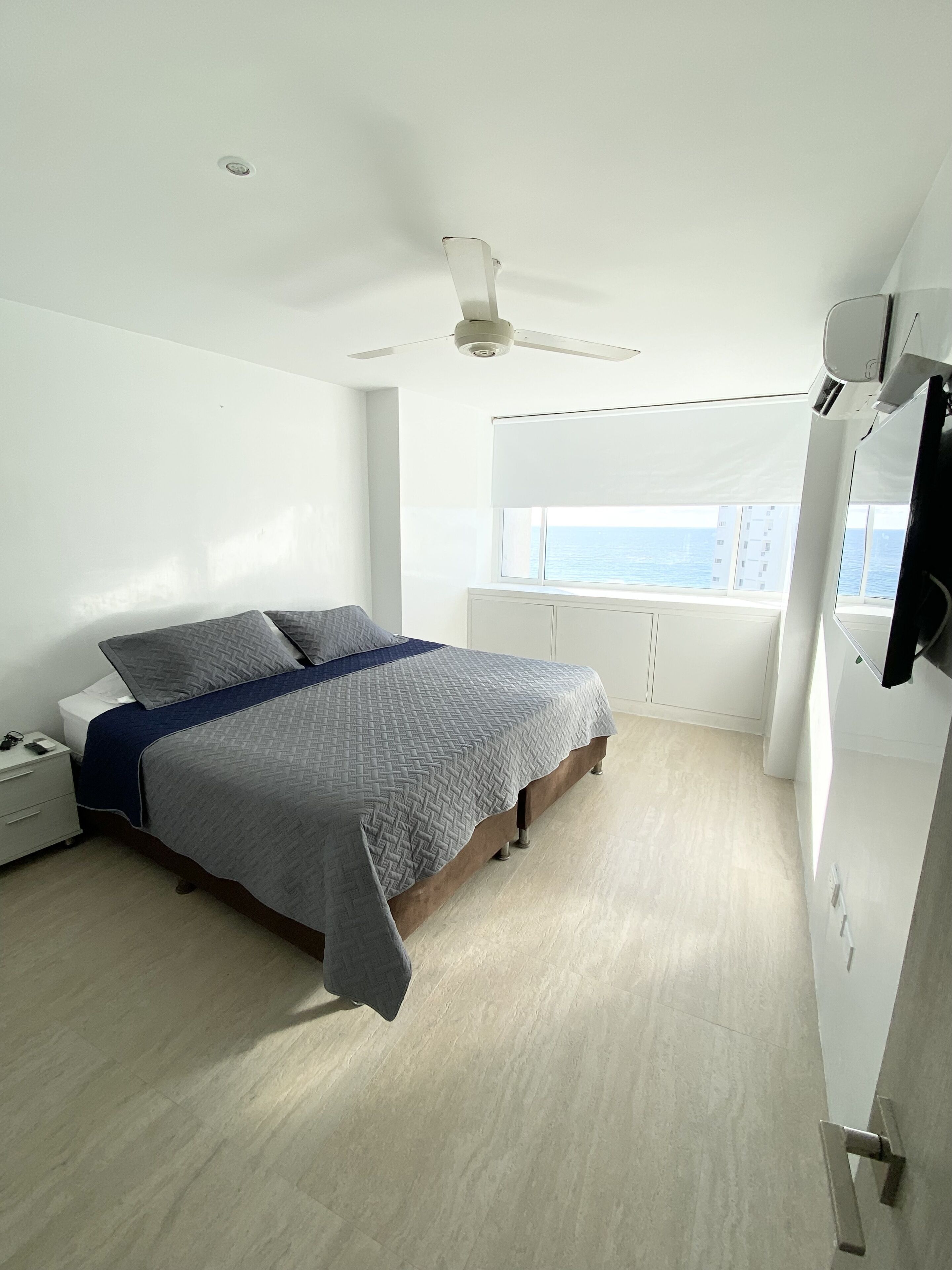 Un dormitorio con cama, mesita de noche, ventana con vista al mar y ventilador de techo.
