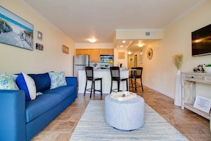 A living room with a blue sofa, a round ottoman, and a kitchen in the background.