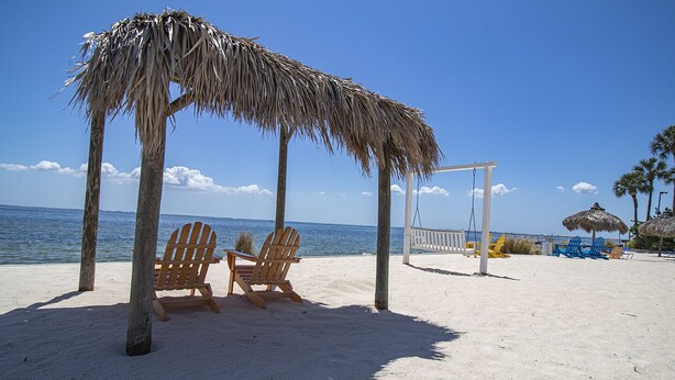 Ein Strand mit Holzstühlen, einer Schaukel und reetgedeckten Sonnenschirmen.