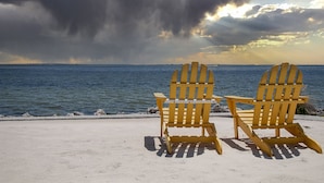 Zwei Adirondack-Stühle blicken auf ein Gewässer, am Himmel sind Wolken.